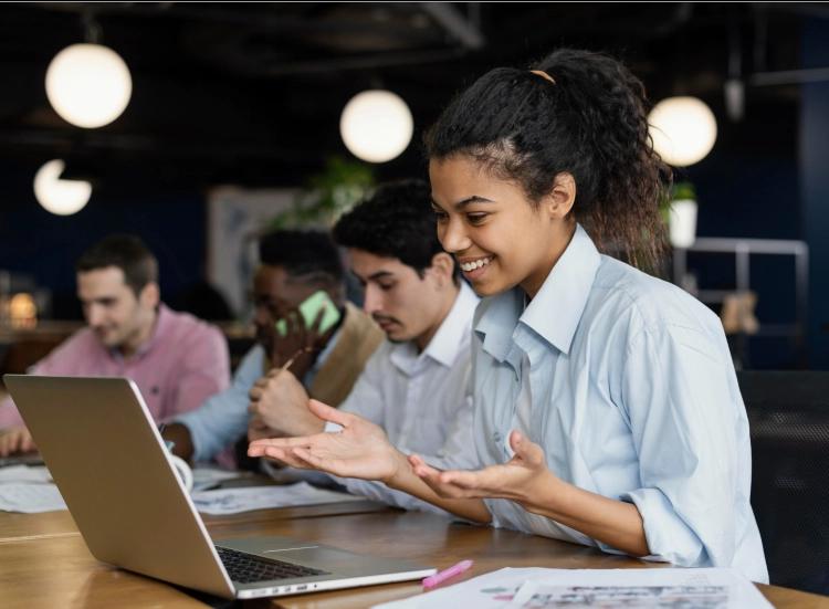 Mulher negra de camisa azul clara sorrindo enquanto gesticula em frente a um notebook. Ao fundo, três pessoas sentadas à mesa, uma falando ao celular e duas analisando papéis.
