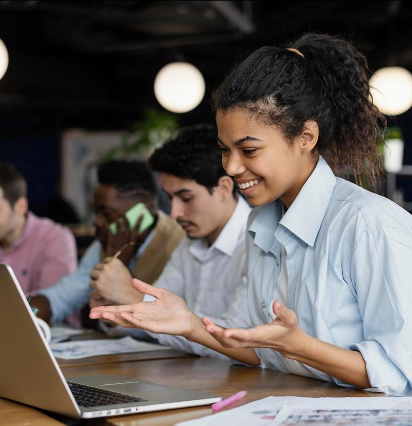 Mulher negra de camisa azul clara sorrindo enquanto gesticula em frente a um notebook. Ao fundo, três pessoas sentadas à mesa, uma falando ao celular e duas analisando papéis.